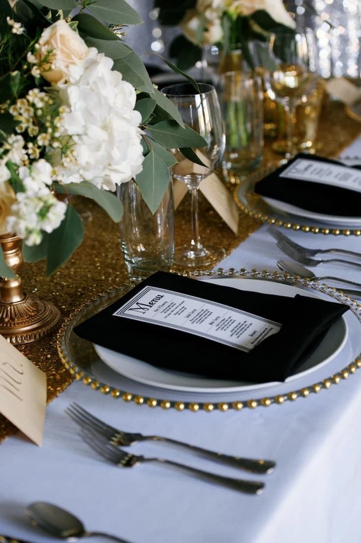 Mise en table élégante avec serviette noire, menu personnalisé et chemin de table à paillettes dorées